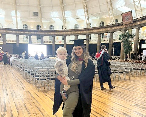 A woman in a graduation gown and cap holds a young child in her arms in a large, empty auditorium.