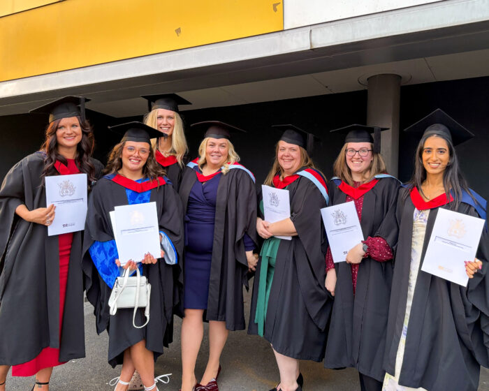 A group of seven women in graduation gowns and caps, holding their diplomas. They are smiling and looking at the camera.