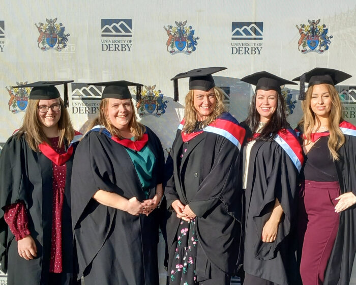 A group of five women in graduation gowns and caps, smiling at the camera. They are standing in front of a brick wall with the University of Derby logo on it.