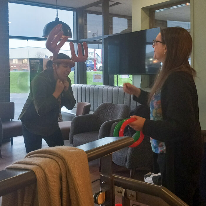 The image shows two people playing a game. The person on the left is wearing a reindeer headband and is trying to catch a ring that the person on the right is throwing. The person on the right is holding a green ring in their hand.