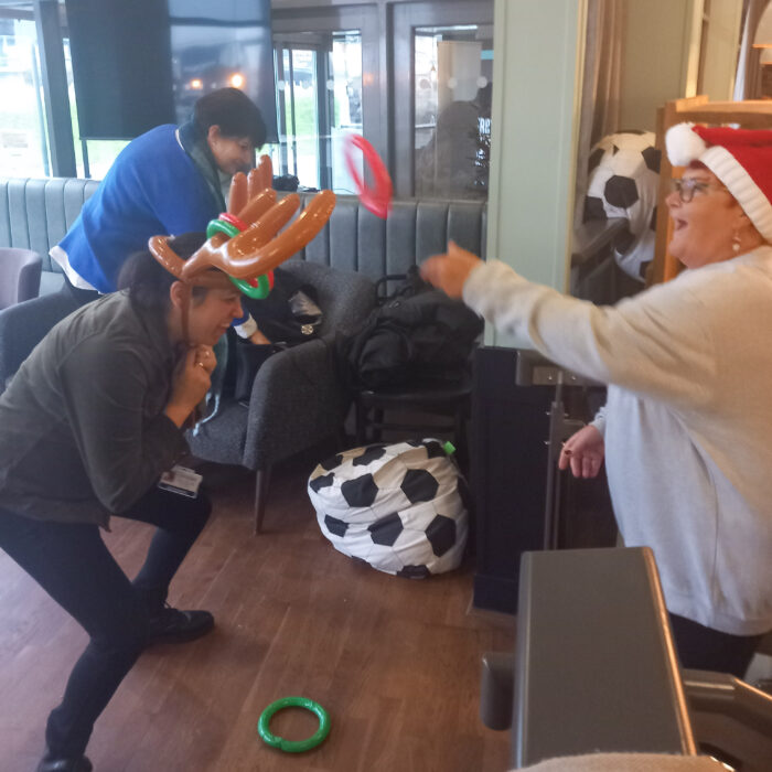 This image shows a group of people having fun at a Christmas party. They are playing a game where they try to throw rings onto a reindeer's antlers. The person in the reindeer antlers is laughing and enjoying the game.