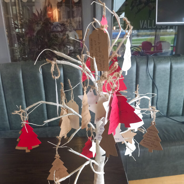 This image shows a Christmas tree made of branches with paper ornaments hanging from it. The ornaments are in the shape of Christmas trees and stars. There are also some handwritten messages on the ornaments.